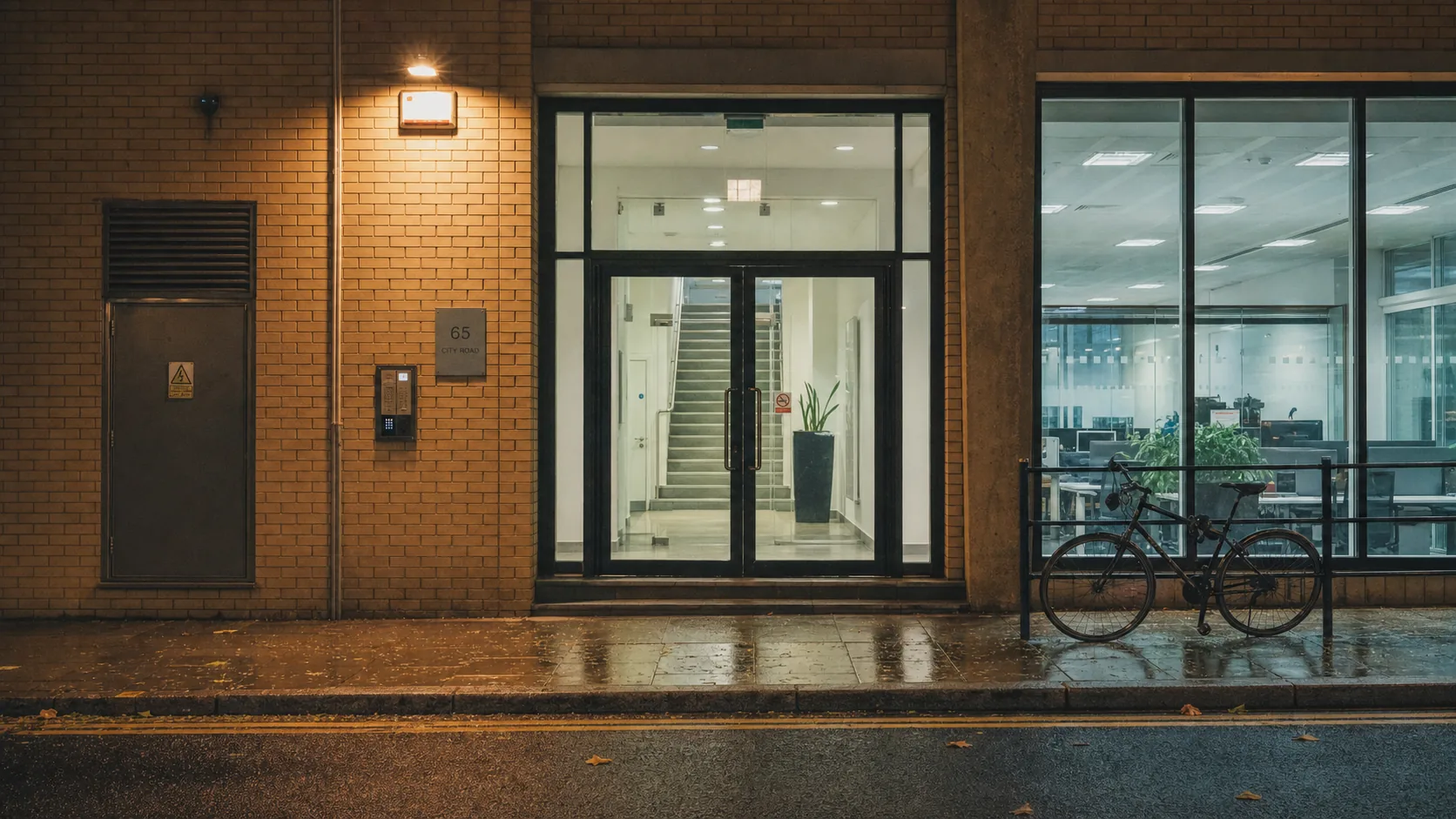A plain London office doorway on City Road at night with an entryphone panel and a lit stairwell visible through the glass.